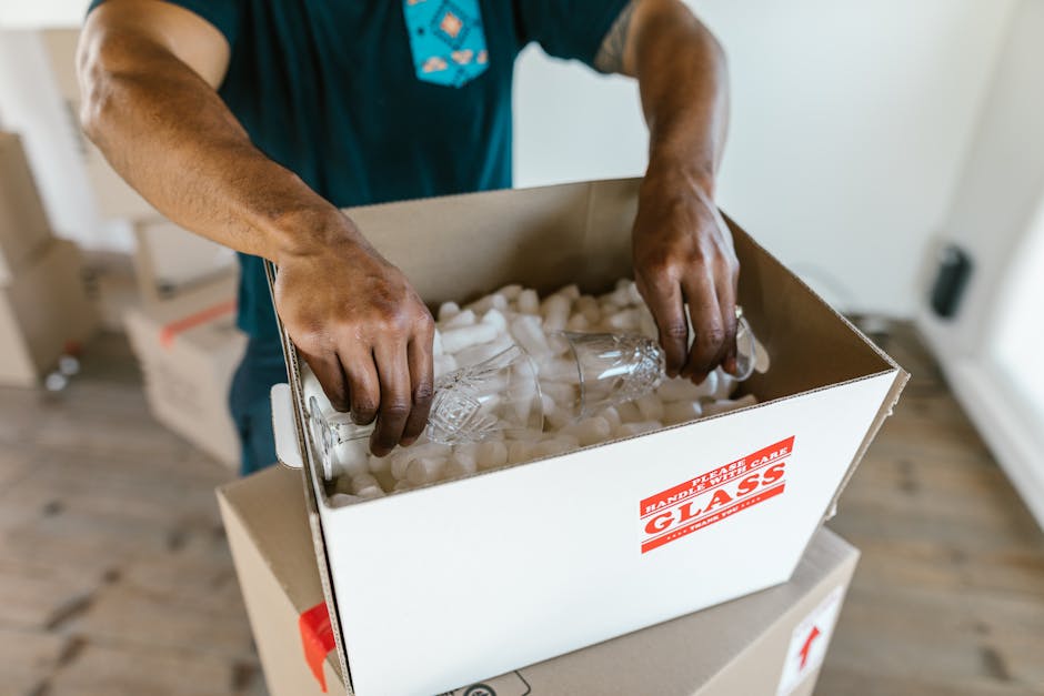 Inside a storage room for home relocation, a person is partially visible behind several cardboard boxes stored on metal shelving. The boxes vary in size, with some marked with a 'fragile' label and a 